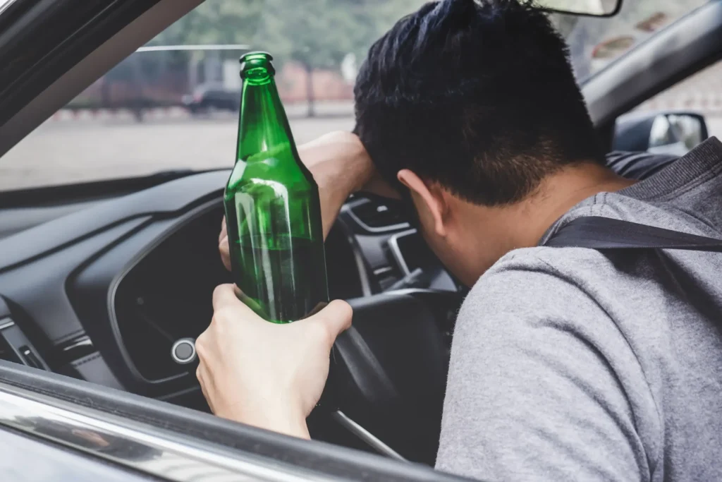 Man slumped over a steering wheel inside a car while holding a green beer bottle, representing the dangers of drunk driving.