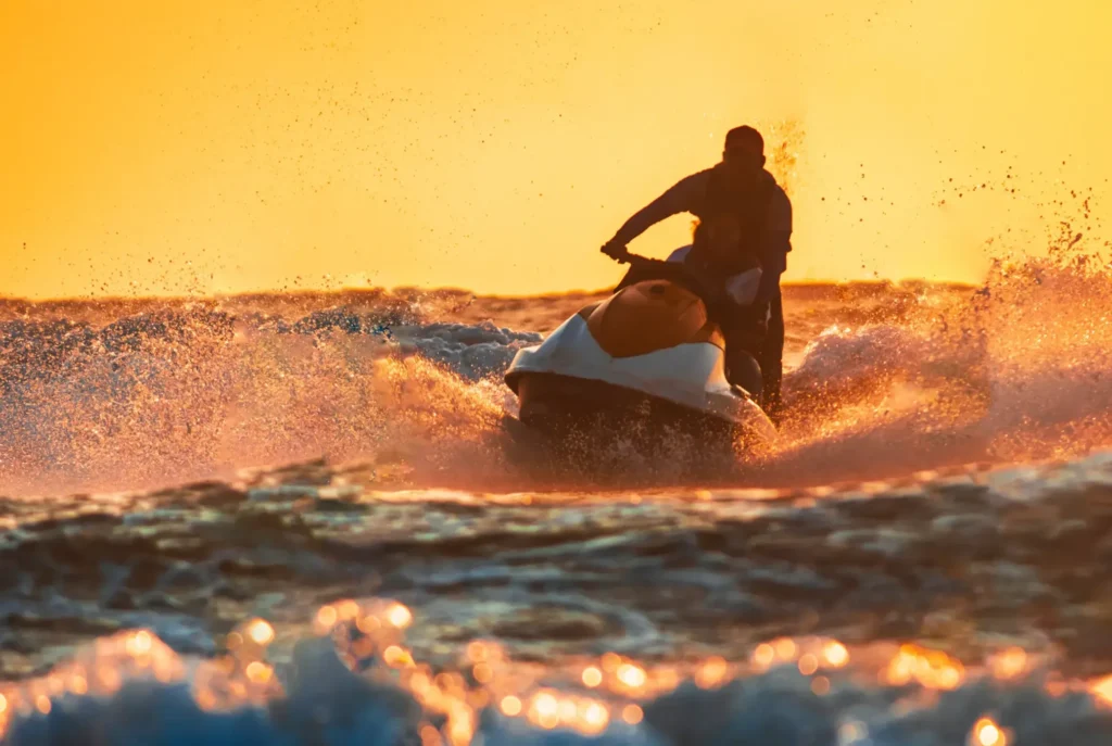 A person riding a jet ski at high speed through ocean waves during a golden sunset, with water spraying.