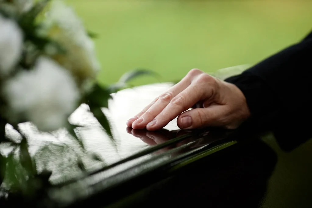 Close-up of a person’s hand resting gently on a polished black casket, with white flowers in soft focus, symbolizing mourning and loss.